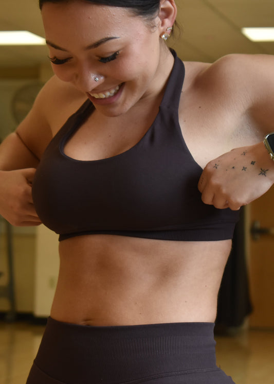 A woman in a gym wearing a dark charcoal gray sports bra and matching high-waisted leggings, adjusting the sports bra. She has a tattoo on her left arm and is wearing a smartwatch.