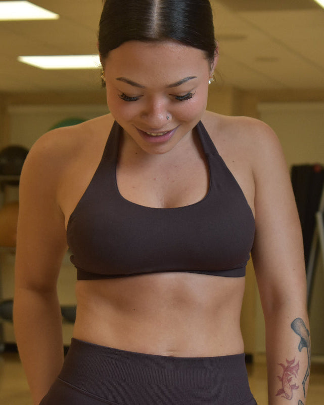 A woman in a gym wearing a dark brown athletic halter sports bra and matching leggings, looking down with a slight smile.